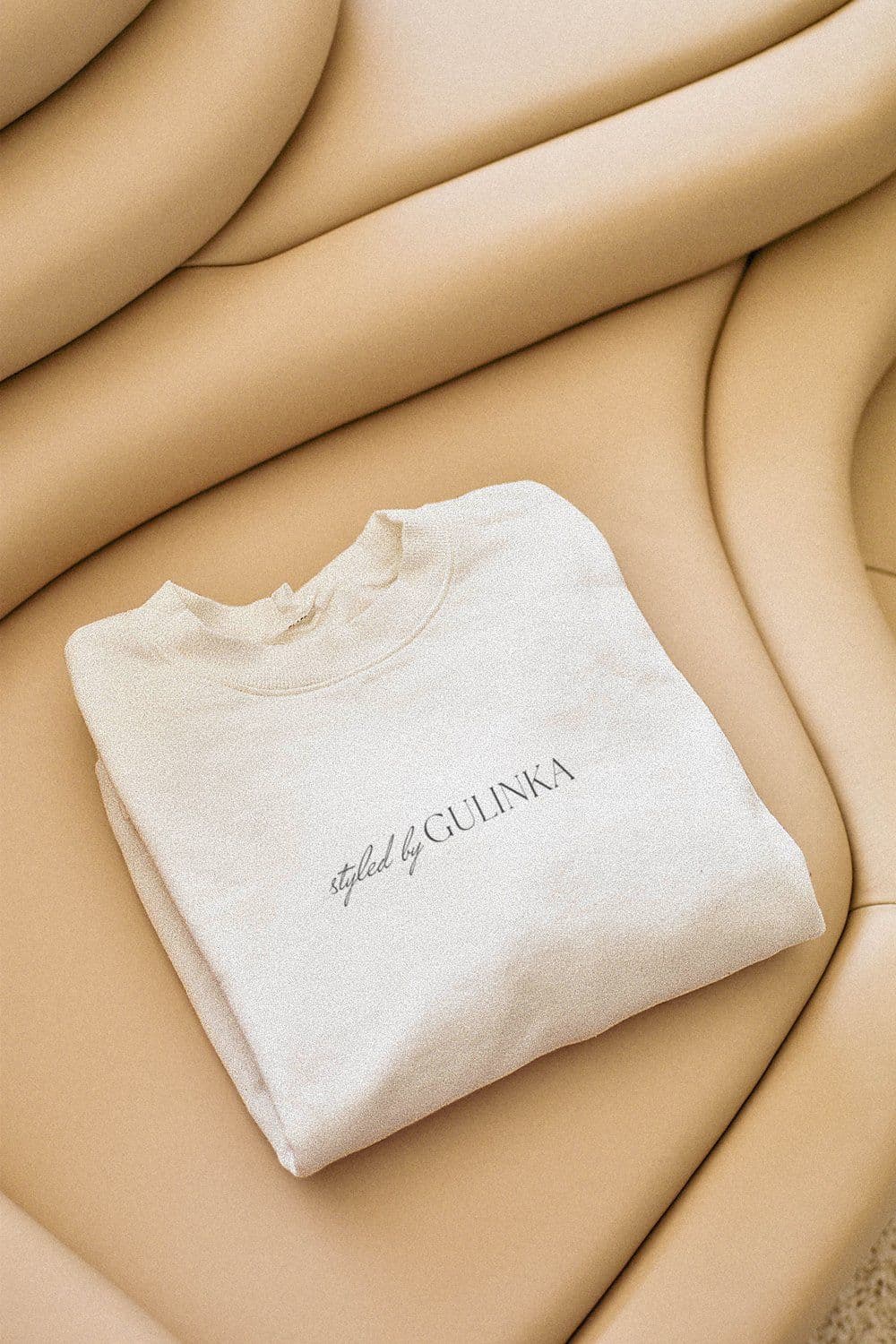 Folded cream sweatshirt with "styled by GULINKA" printed across the chest, resting on a curved beige leather sofa